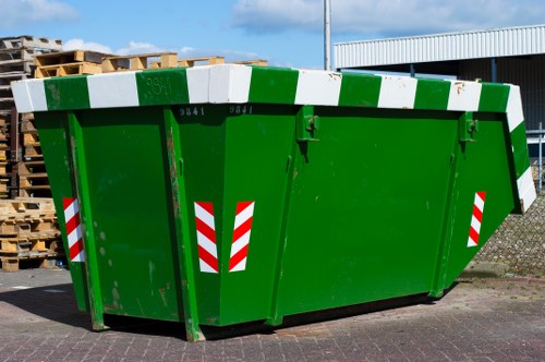 A large green commercial rubbish skip situated on a paved area, positioned outdoors in a warehouse or industrial environment. The skip features a reinforced metal structure with a slightly sloped top edge, painted in a bright green finish with red and white reflective safety markings on the front and sides for visibility. The interior of the skip appears empty or filled with miscellaneous debris not visible in detail. In the background, there are stacks of wooden pallets on the left side and a metal-clad industrial building with a white roof on the right. The sky is partly cloudy, suggesting daylight conditions. The setting indicates a typical waste collection or disposal site, which would be serviced by companies like Commercial Waste Chelsea specializing in rubbish removal services in the Chelsea area. The scene captures the utilitarian nature of waste management infrastructure used for commercial or industrial waste clearance in London, with the skip ready for loading or pickup.