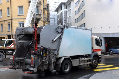 A medium-sized commercial waste collection truck parked on a city street during daylight hours, with a white cabin and a large, rectangular, metallic grey waste container mounted on the back. The container appears to be made of smooth steel with a slightly reflective surface, showing some dirt and minor wear from usage. The truck is positioned near the curb, adjacent to a pedestrian crossing marked with yellow and black stripes on the tarmac. The surrounding environment includes multi-story buildings with a mix of architectural styles, some with yellow facades and others with modern grey finishes. A white ladder extends from the upper part of the truck towards nearby buildings, indicating active rubbish collection or maintenance activity. To the right, a dark grey car is partially visible parked in a parking bay under an overhanging structure, while in the background, additional vehicles and urban fixtures are visible. The overall scene suggests an urban rubbish collection operation, possibly managed by Commercial Waste Chelsea serving the Chelsea area in southwest London, with the streets typical of contemporary city environments.