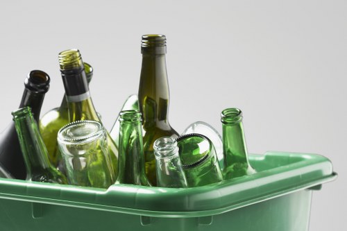 A close-up of a green plastic recycling bin filled with empty glass bottles of various shapes, sizes, and shades of green and brown, with some bottles having caps or rims visible. The bottles are stacked and leaning against each other inside the bin, which is situated on a light grey surface with a plain, neutral background. The scene reflects typical waste collection for glass recycling, commonly provided by rubbish removal services such as Commercial Waste Chelsea, serving areas including Chelsea in London, SW10 postcode. The focus on glass bottles highlights the importance of proper waste segregation and recycling efforts in urban environments to support sustainable disposal practices in the local community.