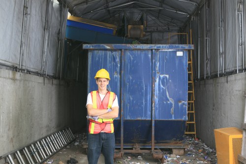 A construction worker wearing a yellow safety helmet, high-visibility vest, and casual clothing stands with arms crossed in front of a large blue industrial skip situated inside a metallic waste storage container. The skip appears to be made of metal with visible wear marks and scratches, and is placed on a concrete or similar durable surface, with some loose debris and rubbish scattered around its base. To the right of the skip, a yellow ladder is leaning against the container's side, and to the far right, a yellow waste bin is partially visible. The interior of the container features corrugated metal walls and a curved roof, enhanced by natural lighting coming from the open entrance at the front. This scene likely depicts a waste management or rubbish collection process, emphasizing the types of equipment used in commercial waste removal services in Chelsea or nearby areas. The environment appears clean and organized, supporting professional rubbish clearance for commercial clients, with a focus on sustainable and efficient waste management practices managed by Commercial Waste Chelsea.