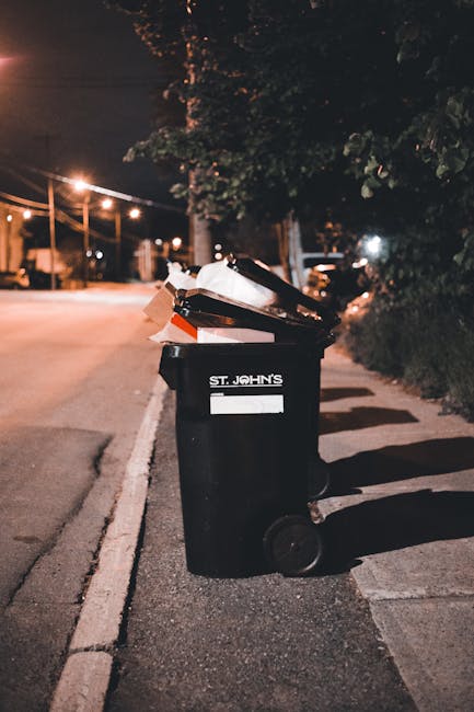 A black wheeled rubbish bin standing on a paved urban sidewalk at night, partially filled with discarded cardboard and paper waste, with white Identification labels and a partial logo reading 'ST. JOH