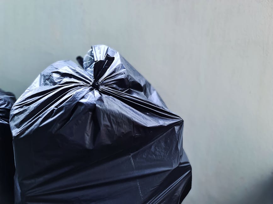 A black wheeled rubbish bin standing on a paved urban sidewalk at night, partially filled with discarded cardboard and paper waste, with white Identification labels and a partial logo reading 'ST. JOH