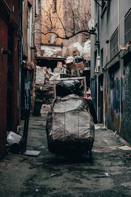 A white waste collection truck with an open rear compactor, parked on a narrow cobblestone street in an urban residential area. The truck’s rear is rusted and dirty, with visible mechanical componen