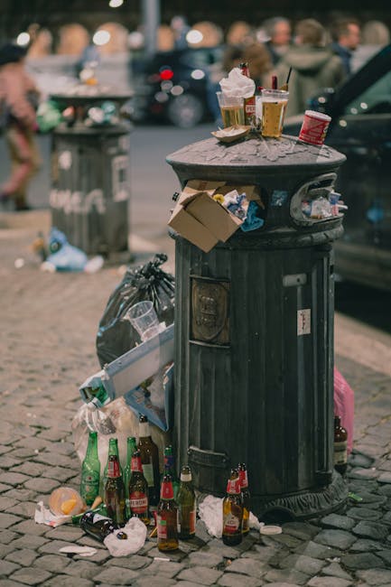 An outdoor scene showing a pile of mixed waste debris on a gravel surface, including black plastic rubbish bags, a large yellow plastic tray, and a visibly worn, dirty beige car tire discarded among t