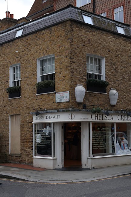 A three-story corner building constructed of brown brick with a slightly weathered texture stands on a street corner, featuring a ground-floor shop with a white fascia displaying the name 'Chelsea Gre